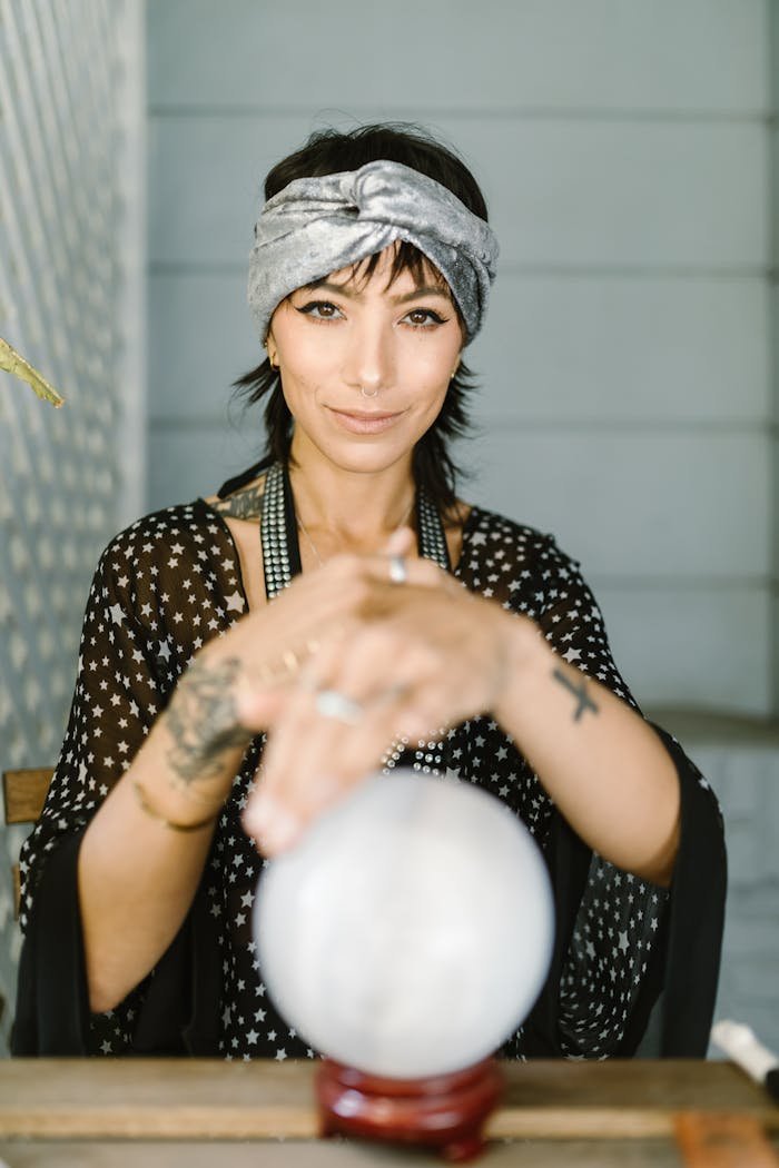 A woman with a headscarf and tattoos gazes over a crystal ball in a mystical setting.