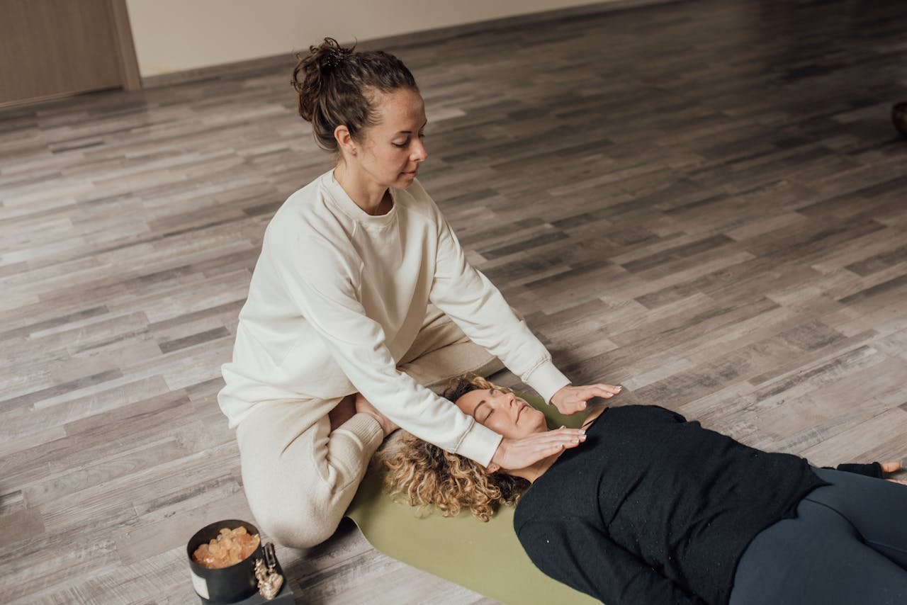 Woman practicing Reiki healing therapy on another woman lying indoors on a yoga mat.